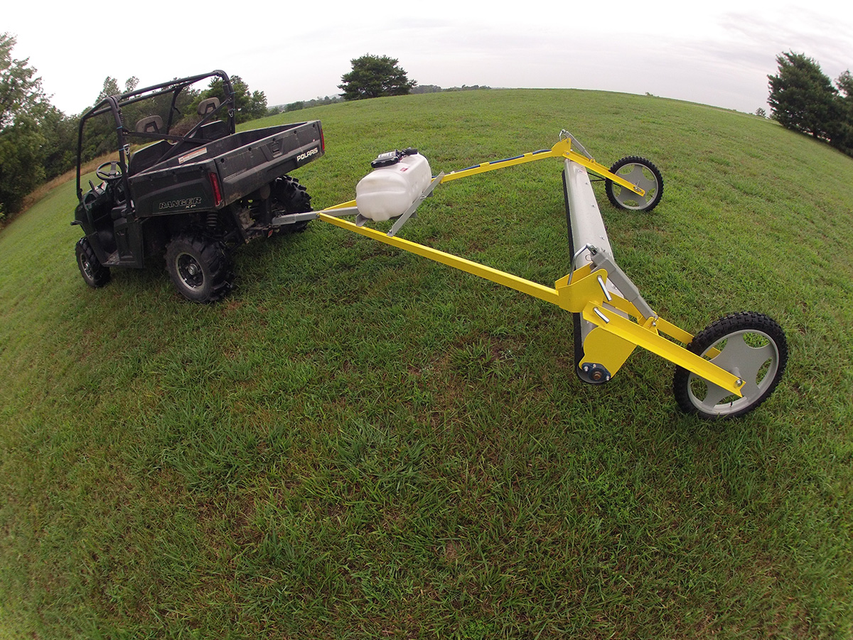 GrassWorks Pull-Type Weed Wiper™ connected to an ATV, treating weeds in an open field.