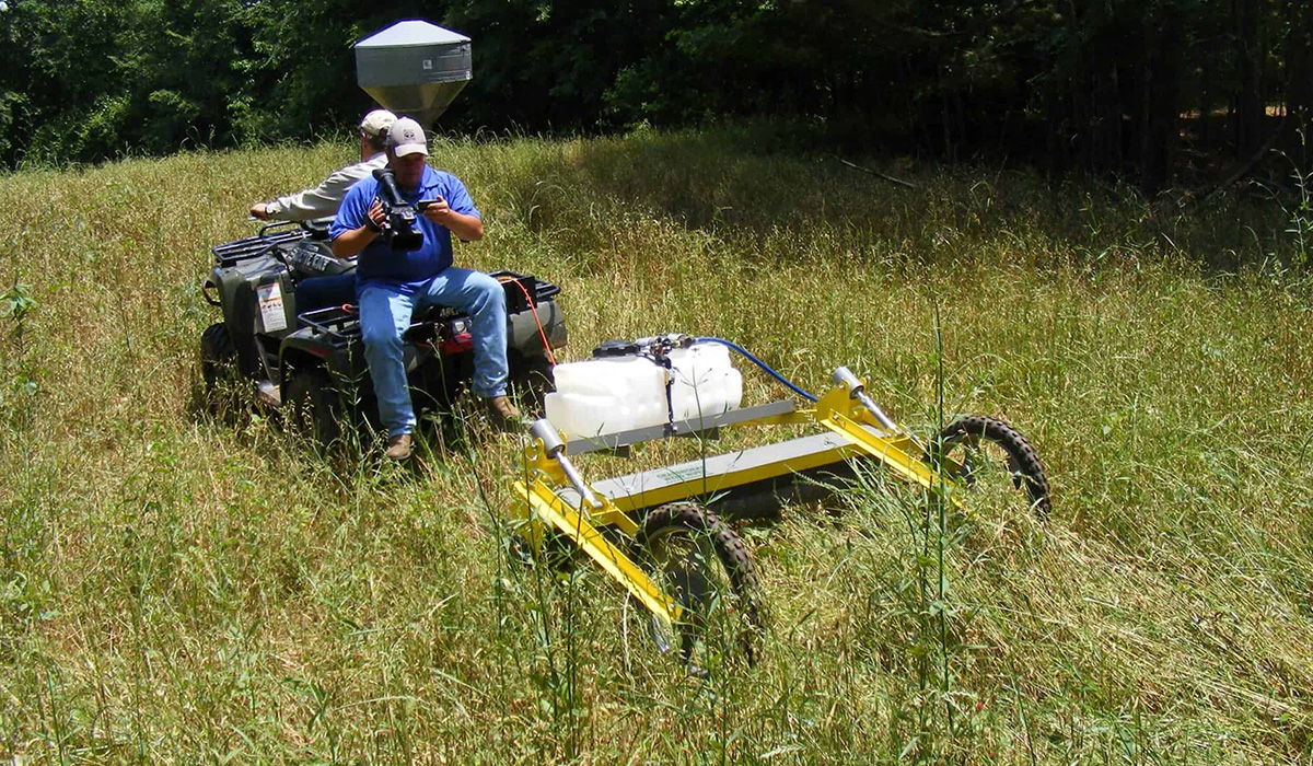 Two-person ATV operating a GrassWorks Pull-Type Weed Wiper™ in tall grass.