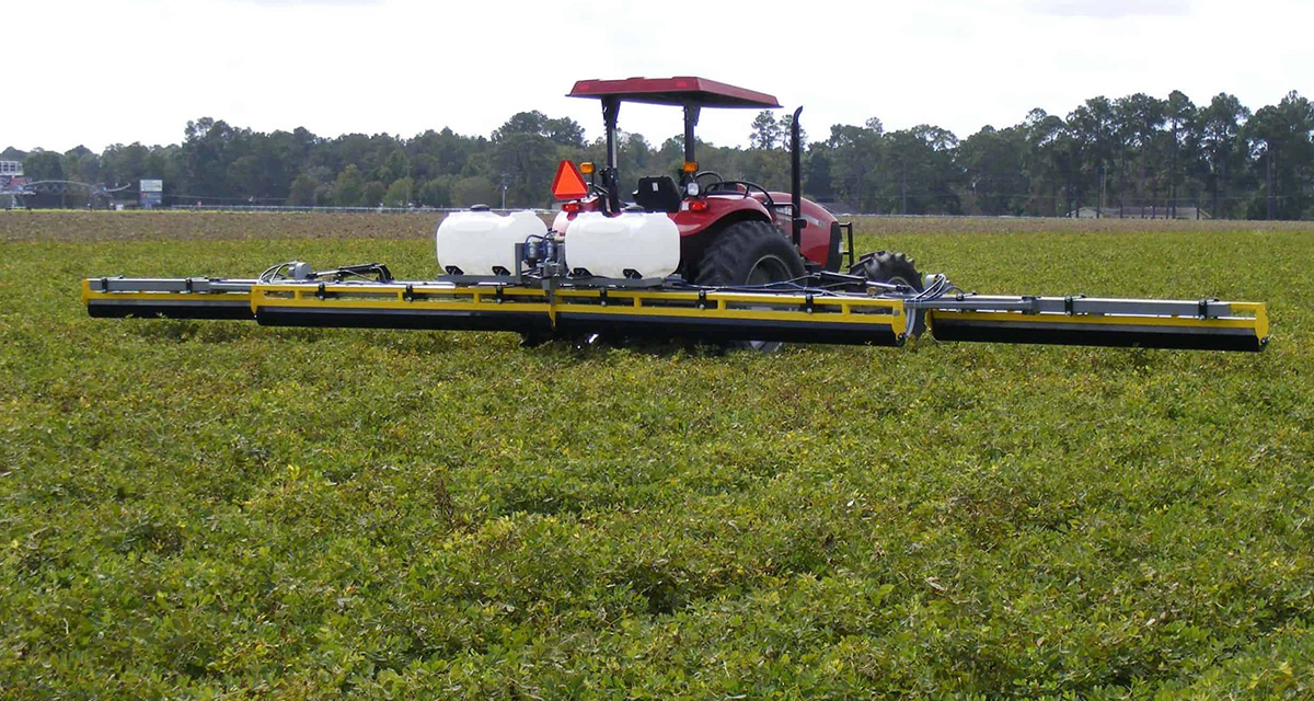 A GrassWorks Manufacturing Weed Wiper mounted on a red tractor, treating vegetation in an open field.