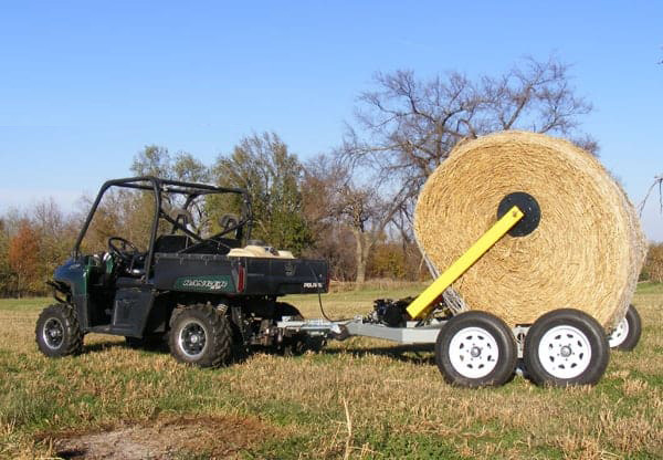 Hydraulic hay bale handler attached to an ATV, actively unrolling a round bale in a pasture, reducing hay waste and optimizing livestock feeding.